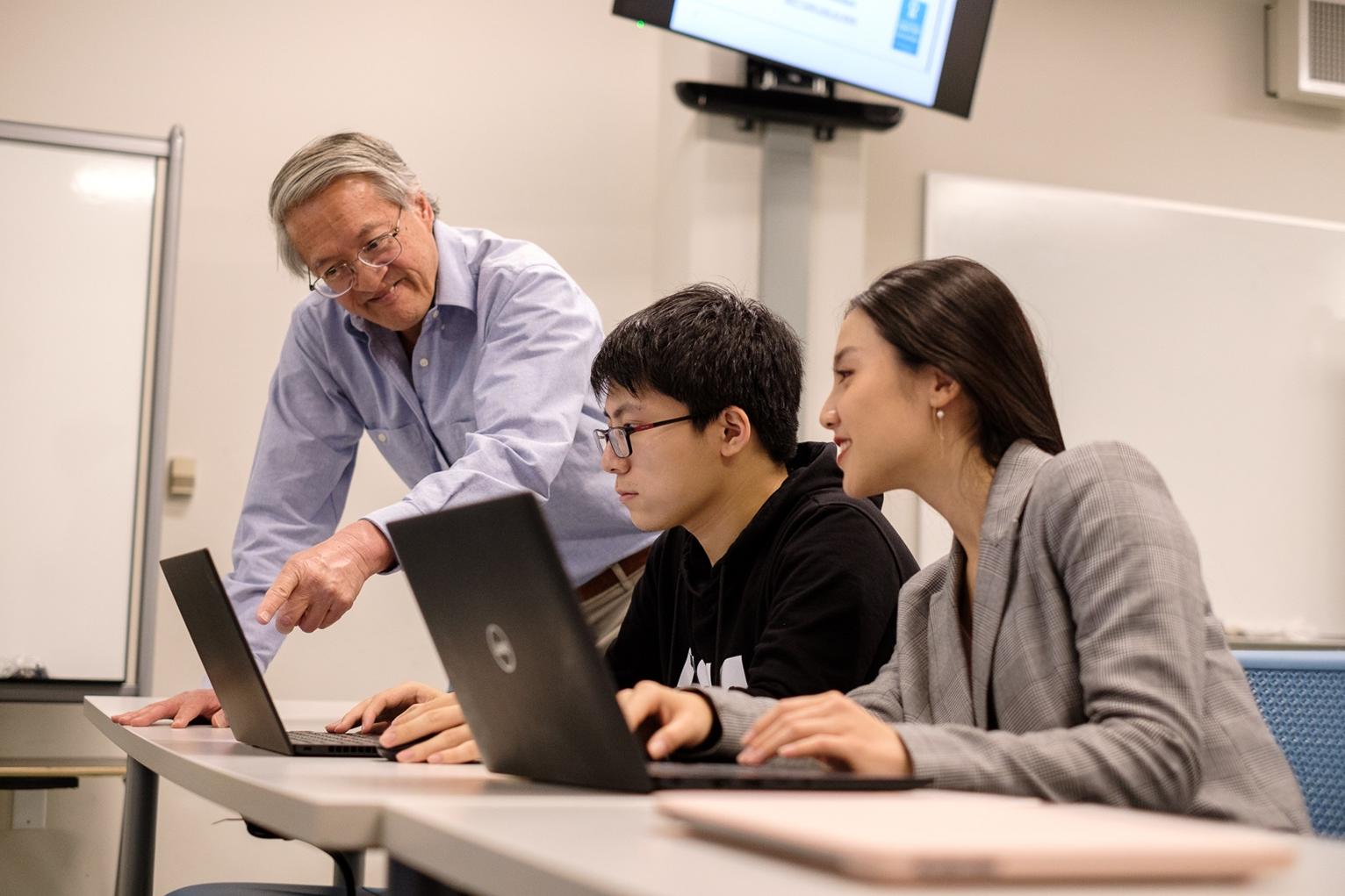 Two students sit at a desk in front of a computer with a professor teaching them at 菠菜网lol正规平台. 